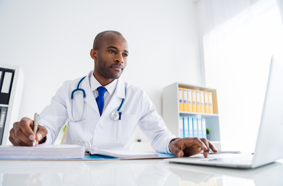 physician sitting at his desk and completing physician training on a computer and writing in his notebook